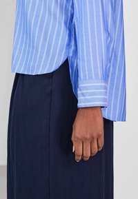 Light blue striped shirt with a curved hem, long sleeves, and a buttoned cuff, paired with a dark navy skirt. Smooth fabric texture.