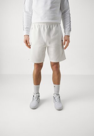 Mann trägt weiße Sportshorts, weißes Langarmshirt mit Streifen an den Ärmeln, weiße Socken und weiße Sneaker, steht vor weißem Hintergrund.
