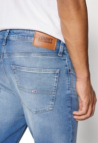 Ljust blå denimjeans med en märkt brun läderpatch som står "TOMMY JEANS." Funktioner inkluderar vinklade bakfickor och detaljerad sömnad.