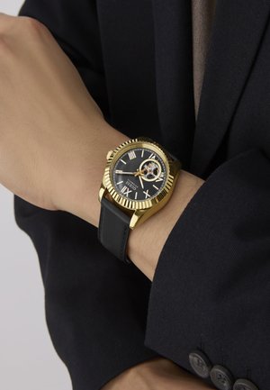 Gold-tone watch with a black dial, Roman numerals, and clear case back. Features a black leather strap and textured bezel, worn on a wrist.