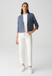 Blazer cropped bleu avec deux boutons, manches longues, en matière texturée ; associé à un pantalon blanc à jambe droite et des baskets blanches.