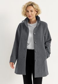 Grau-wollmischung Mantel mit hohem Kragen, sechs Knöpfen und zwei Seitentaschen; lässige Passform und weiche Textur; getragen über einem weißen Hemd.