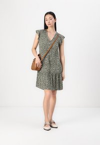 Zelené květinové šaty s výstřihem do V, s volánovými rukávy a vrstevnatým lemem. V kombinaci s plochými, světlými botami a hnědou crossbody taškou.