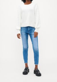 Biała sweter z teksturowanymi, bufkowymi rękawami i małymi ozdobnymi koralikami, zestawiony z jasnoniebieskimi obcisłymi dżinsami i masywnymi czarnymi platformami.