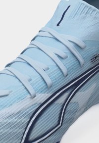 Približen posnetek svetlo modrega športnega čevlja z belimi vezalkami in temno modrim geometričnim logotipom na teksturiranem tkaninskem zgornjem delu.