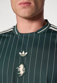 Grønt stribet atletisk skjorte med rund hals, der har et broderet logo og et dekorativt heste grafik på forsiden. Hvide accenter ved skuldrene.