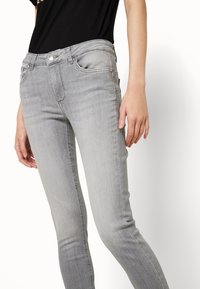 Vaqueros pitillo de mezclilla gris con cintura de tiro medio, bolsillos delanteros y sutiles detalles de desgaste. Textura suave con un diseño ajustado al cuerpo.