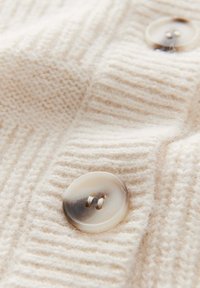 Cremefarbenes Strickmaterial mit gerippter Textur, das einen großen, runden Zweilochknopf in einem hellen, marmorierten Muster zeigt.