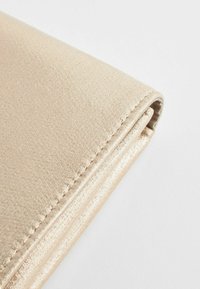 Pochette beige métallique pliée avec coutures visibles sur tissu lisse texturé, sur un fond clair uni.