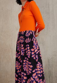 Kvinde iført en lys orange langærmet top med lynlås og sort nederdel med høj talje med orange og lilla bladmønster, stående mod en ensfarvet væg.