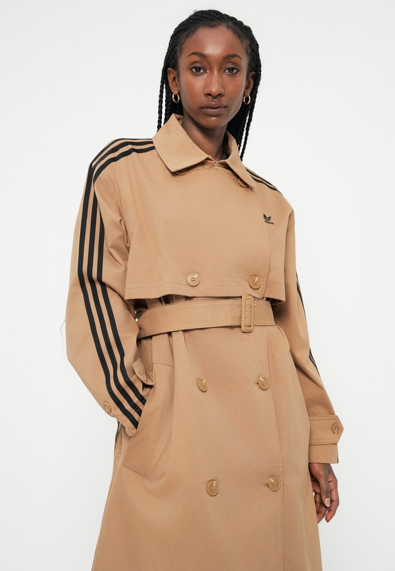 Tan dobbelbrystet trenchcoat med sorte striber på ærmerne, bælte i taljen, knapdetaljer og en klassisk krave. Glat stofstruktur.