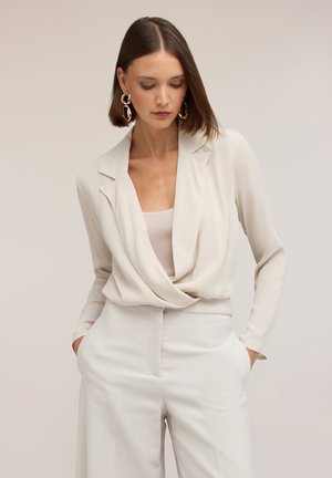 Mujer con cabello castaño liso lleva pendientes grandes de aro dorados, un blazer crema drapeado y pantalones de talle alto a juego con las manos en los bolsillos.