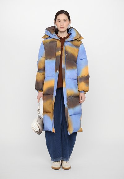 Gepolsterter Mantel mit einem blauen, braunen und gelben Farbverlauf-Design. Mit Kapuze, Druckknopfverschluss vorne und Seitentaschen. Kombiniert mit weit geschnittenen Jeans und weißen Schuhen.
