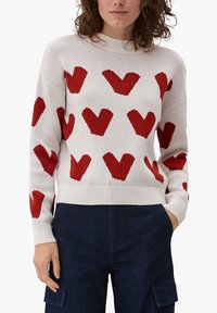 Maglione a maglia color crema con grandi motivi a cuore rosso, scollo rotondo e polsini a coste. Abbinato a pantaloni cargo blu scuro.