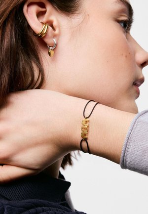 Close-up of a person’s wrist with a black cord bracelet spelling "MOM" in gold and ear with gold and silver hoop earrings with a heart charm.