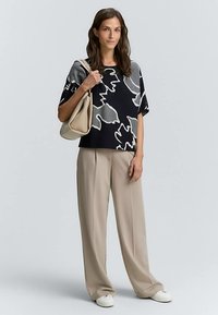 Zwart en grijs top met abstract patroon en losse mouwen, gecombineerd met beige wijde broekspijpen en witte sneakers, terwijl ze een beige handtas vasthoudt.