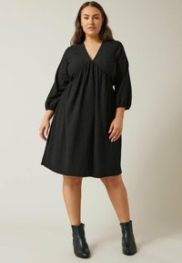 Schwarzes knielanges Kleid mit V-Ausschnitt, langen puffigen Ärmeln und gerafftem Taillendetail. Hergestellt aus einem strukturierten Stoff. Getragen mit schwarzen Stiefeletten.