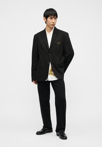 Άνδρας που φοράει μαύρο σακάκι με δύο κουμπιά, λευκό πουκάμισο με χρυσό σχέδιο, μαύρο παντελόνι και μαύρα παπούτσια, όρθιος μπροστά σε απλό ανοιχτό φόντο.