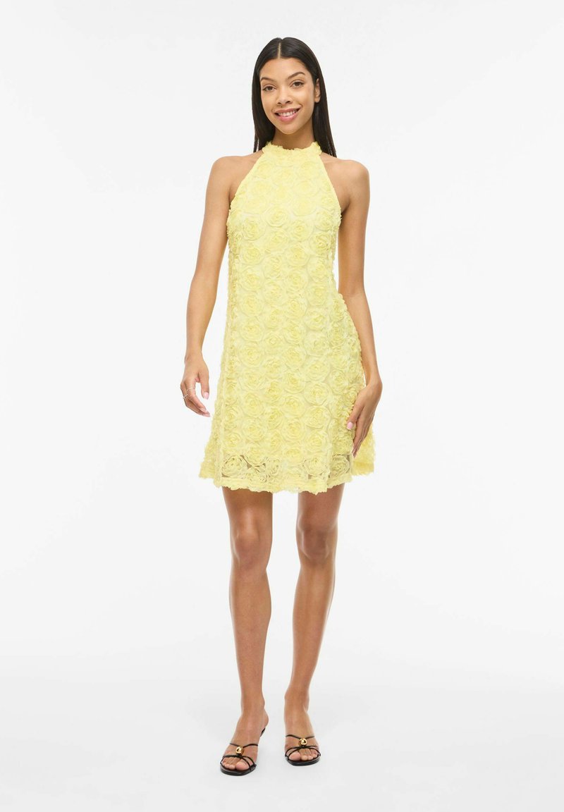 Femme souriante portant une robe jaune sans manches en dentelle avec texture florale et des talons noirs ouverts, se tenant devant un fond blanc.