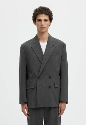 Jeune homme aux cheveux bouclés portant une veste de costume double boutonnage à fines rayures gris foncé oversize et un pantalon assorti sur une chemise blanche.