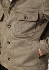 Veste en daim vert olive avec deux poches poitrine, fermeture à boutons et texture lisse. Coupe ajustée avec poches latérales.
