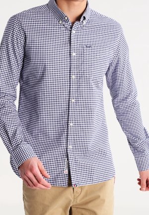 Mann trägt ein langärmliges, geknöpftes Hemd mit marineblau-weiß kariertem Muster und beige Hosen, steht vor einem schlichten Hintergrund.