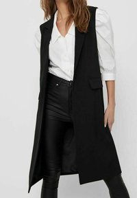 Czarna bezrękawnik wykonana z gładkiego materiału o dopasowanym kroju, z bocznymi kieszeniami i klapami w kształcie notcha, w zestawie z białą koszulą.