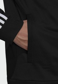 Chaqueta deportiva negra con tejido texturizado, que presenta un bolsillo lateral y franjas blancas en contraste en la manga. Primer plano de una mano en el bolsillo.