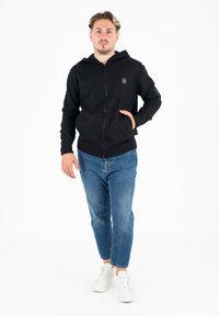 Felpa nera con zip e tasca frontale, polsini a costine, abbinata a jeans azzurri chiari e sneakers bianche su uno sfondo semplice.