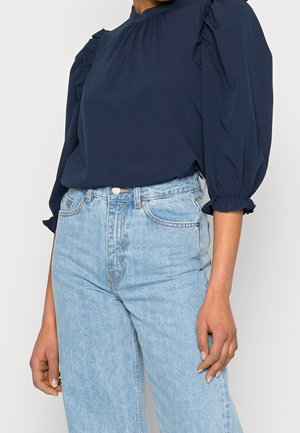 Vrouw draagt een marineblauw blouse met pofmouwen, ingestopt in een lichtblauwe spijkerbroek met hoge taille, staand met ontspannen armen langs het lichaam.