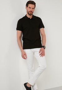 Černé polo tričko s krátkými rukávy, spárované s bílými slim kalhotami a černými teniskami. Hodinky na zápěstí, postávající proti bílé zdi.
