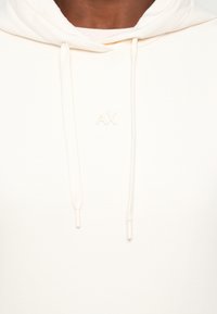 Gros plan d'une personne portant un sweat à capuche blanc avec une capuche et des cordons, avec un logo embossé "AX" sur la zone de la poitrine.