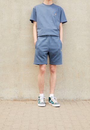 Blue cotton t-shirt and matching shorts, both with a relaxed fit. Notable gold logo on the chest. Footwear: teal and gray high-top sneakers.