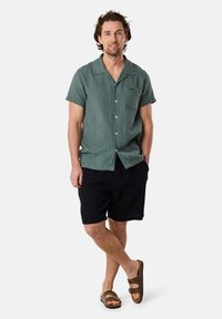 Maglietta verde a maniche corte con tessuto strutturato, pantaloncini neri a coste e sandali marroni. La maglietta presenta una tasca e bottoni; vestibilità comoda complessiva.