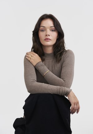 Young woman with dark hair wearing a brown long-sleeve top and black skirt, sitting with arms crossed, wearing gold rings and bracelet.