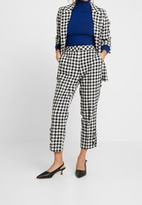 Czarnobiała kratka w stylu gingham na garniturze z dopasowanym marynarką i spodniami z wysokim stanem. Pod spodem niebieski golf. Czarne, szpiczaste buty na obcasie z paskiem z tyłu.