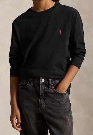 Personne portant un t-shirt noir à manches longues avec un petit logo rouge sur la poitrine gauche et un jean gris foncé, une main dans la poche.