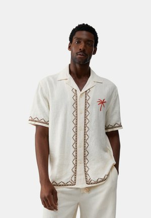 Uomo che indossa una camicia crema a maniche corte con bottoni, con bordo geometrico marrone e un piccolo ricamo di palma rossa sul petto.