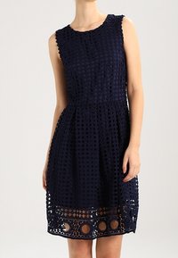Námořnická modrá bezramínková šaty s texturovanou krajkovou vrstvou, obsahující diamantové a kruhové výřezy, rozšířenou sukni a přiléhavý pas.