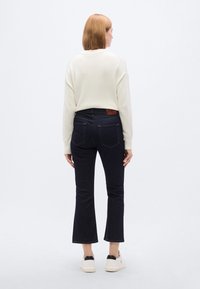 Σκούρο μπλε τζιν με καμπάνα και μεσαία μέση, με καφέ δερμάτινο ταμπελάκι στη μέση πίσω και αντίθετες ραφές. Φοράει λευκό πουλόβερ.