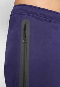 Lila sportnadrág, oldalsó varrással és fekete cipzáras zsebbel. Sima textúra, a derékrészen rugalmas gumival.