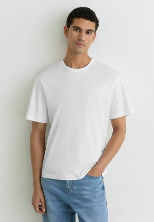 Jonge man met kort krullend haar, gekleed in een eenvoudig wit T-shirt met korte mouwen en een blauwe spijkerbroek, staand tegen een witte muur.