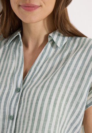Kvinde iført en grøn og hvid vertikalt stribet skjorte med knapper og krave, vist fra hagen til midten af overkroppen mod en lys baggrund.