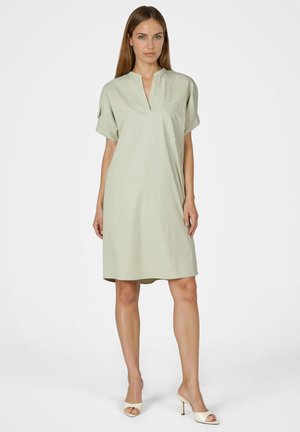 Frau steht und trägt ein hellgrünes, knielanges Kleid mit kurzen Ärmeln, V-Ausschnitt und Tasche, kombiniert mit weißen Sandalen mit Absatz.