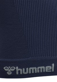 Gros plan sur un tissu en maille côtelée bleu marine avec le logo gris de la marque hummel et l'emblème d'abeille tissés près du bord inférieur.