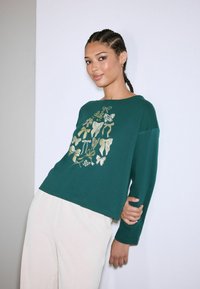 Zielona bluzka z długim rękawem w złoty kwiatowy wzór z kokardą, zestawiona z jasnymi, strukturalnymi spodniami o szerokiej nogawce. Miękka tkanina, luźny krój.