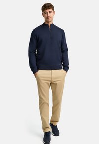 Navyblauer Zip-Up-Pullover mit geripptem Kragen, kombiniert mit beigen Chinos und dunklen Sneakers, steht vor einem schlichten weißen Hintergrund.