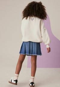 Crème sweatshirt met "L.A. 1982" op de achterkant, gecombineerd met een blauwe geplooide denim rok en zwart-witte sneakers. Getextureerde, ontspannen pasvorm.