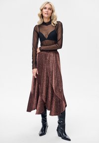 Czarna przezroczysta bluzka z długim rękawem, noszona na brązowej błyszczącej, rozszerzanej długiej spódnicy, zestawiona z czarnymi kozakami do kolan.