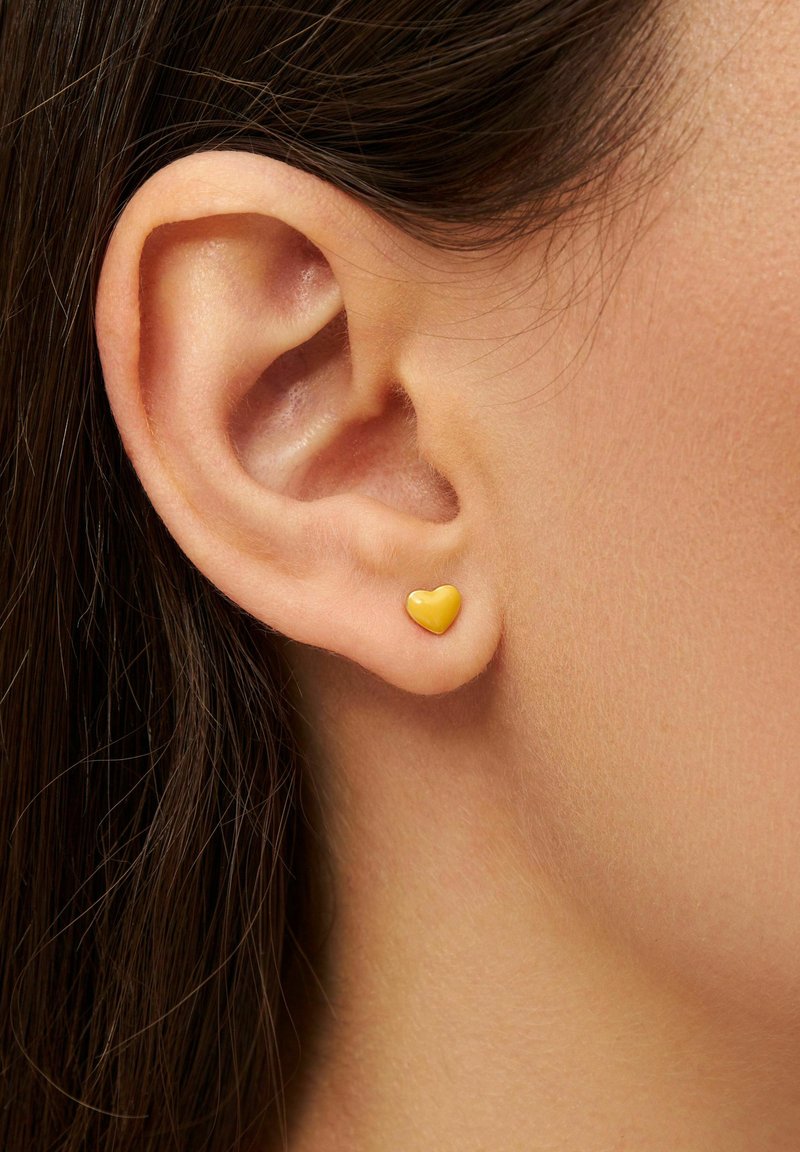 Gul hjärtformad studsörhänge, tillverkat av glänsande material, visat mot huden med delvis synligt hår. Enkel och minimalistisk design.